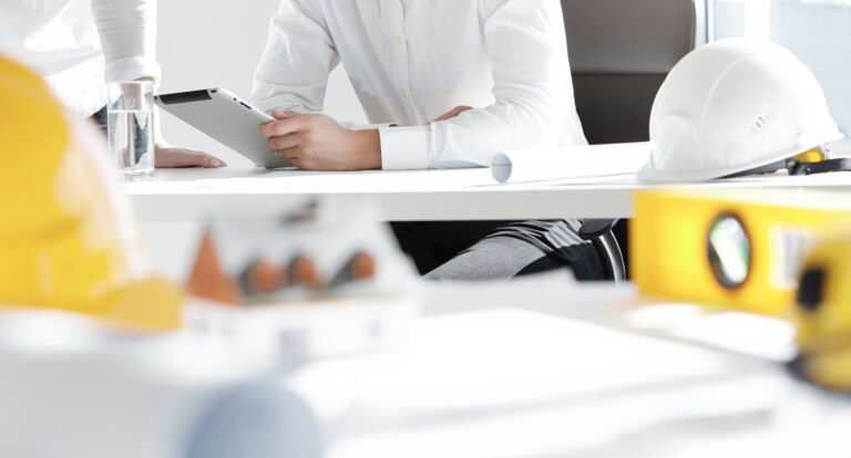 two engineers or designers working on construction project, analyzing blueprint on digital tablet, discussing plans in office with scale model house, helmets and architectural tools in foreground